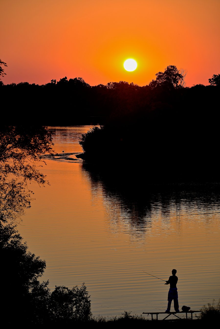 A man stands on a picnic table while fishing as the sun hovers just above the horizon.