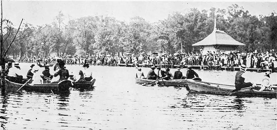 People row across a lagoon with hundreds of others standing on a bank.