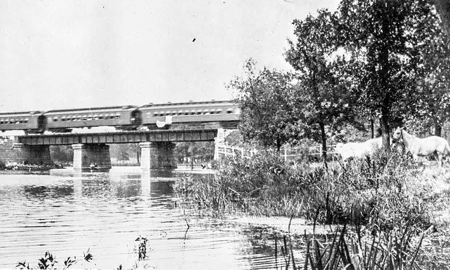 A train crosses a bridge above a river in an old black and white photograph.