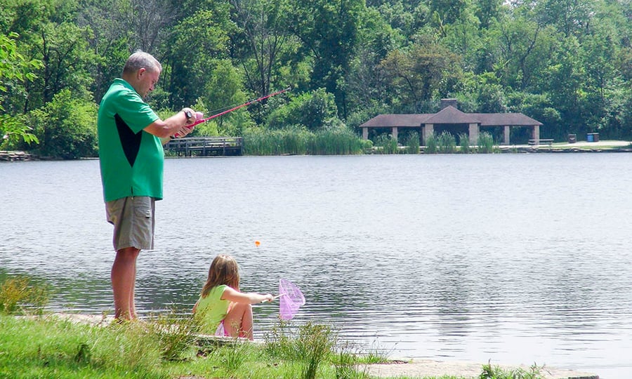 On a lakeshore a man holds a fishing pole while his daughter gets close to the water with a net.