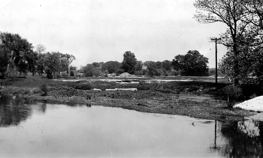 A black and white photo showing a shoreline with a mound and trees in the distance.
