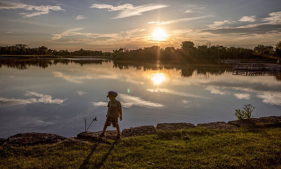 A boy stands on the shoreline of a lake while the sun hovers near the horizon.