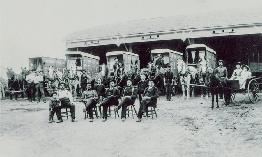 A black and white photograph shows men sitting in chairs in front of horse-drawn wagons.