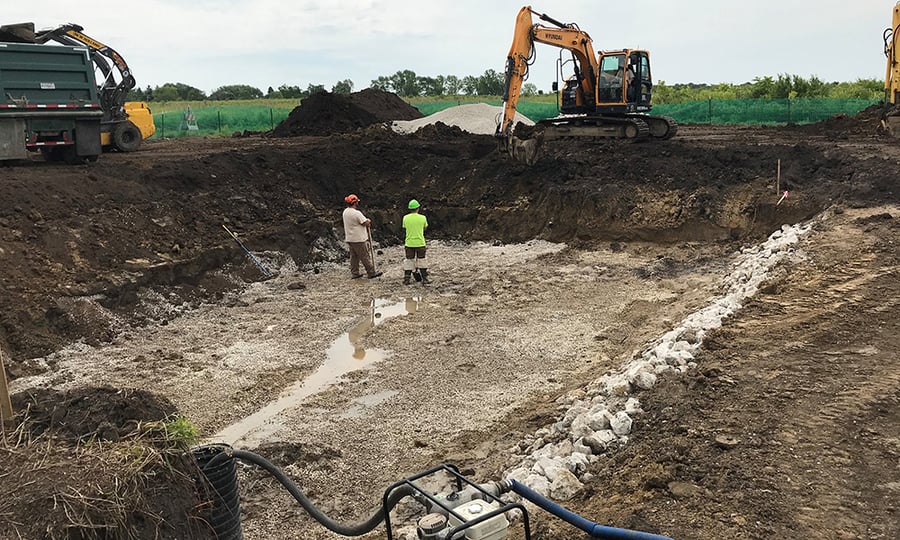 Two men in hard hats stand in a ditch being worked on with heavy machinery.