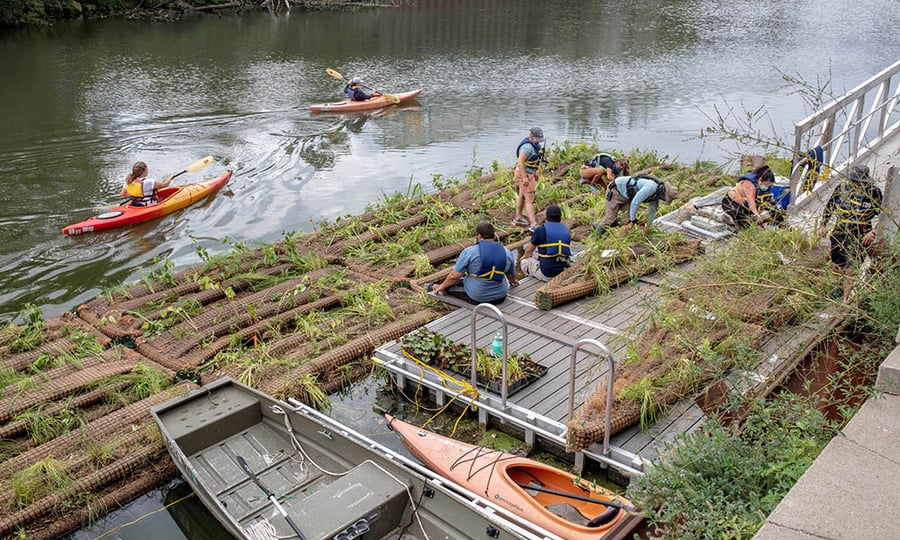Two kayakers paddle past a floating wetland that is being worked on by about eight people.
