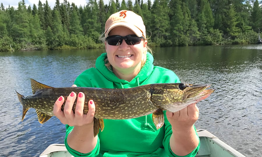 A woman holds a northern pike while in a boat on a lake.