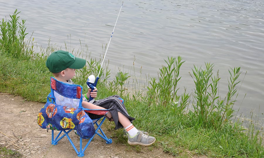 A boy fishes from a foldable chair on a lakeside trail.