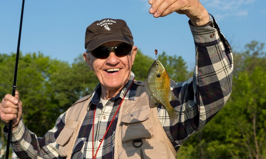 A man in a hat, sunglasses, and canvas vest smiles as he holds up a sunfish with a hook in its mouth.