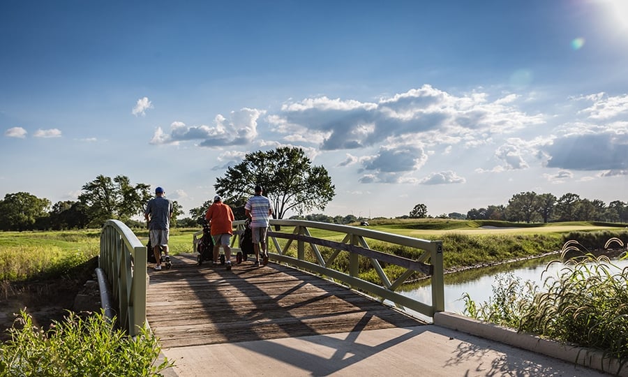 Three golfers with push carts cross a bridge at a course.