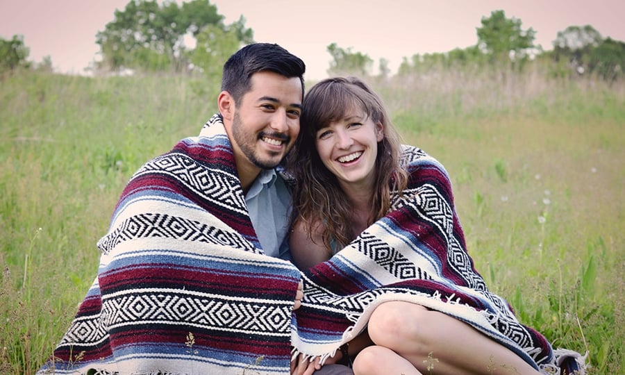 A couple sits, smiling in a field wrapped in a blanket.