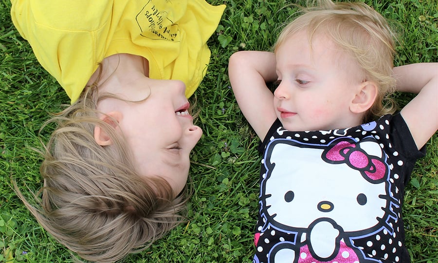 Two children lie in a grassy field smiling at one another.
