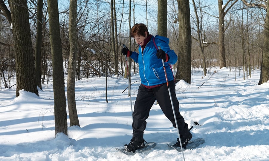 A woman in a blue jacket and black pants snow shoes through a forest.