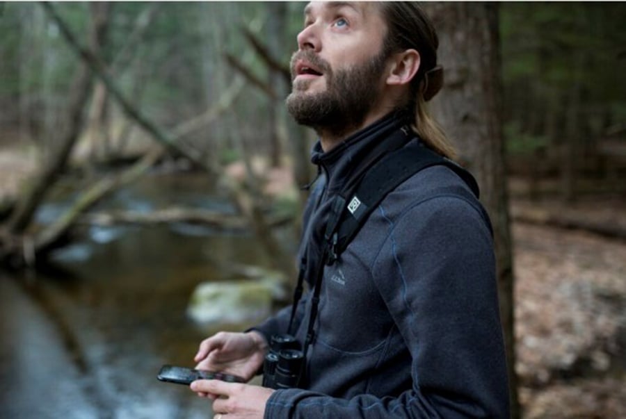 A man with binoculars around his neck looks up for birds in a forest.