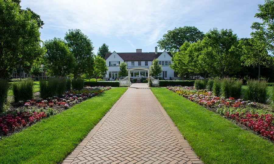A brick walk lined by flowers leads to a white house.