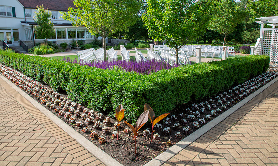White chairs in a courtyard are surrounded by well-manicured bushes and flowers.
