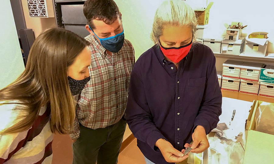 Three people in facemasks look at an old arrowhead.