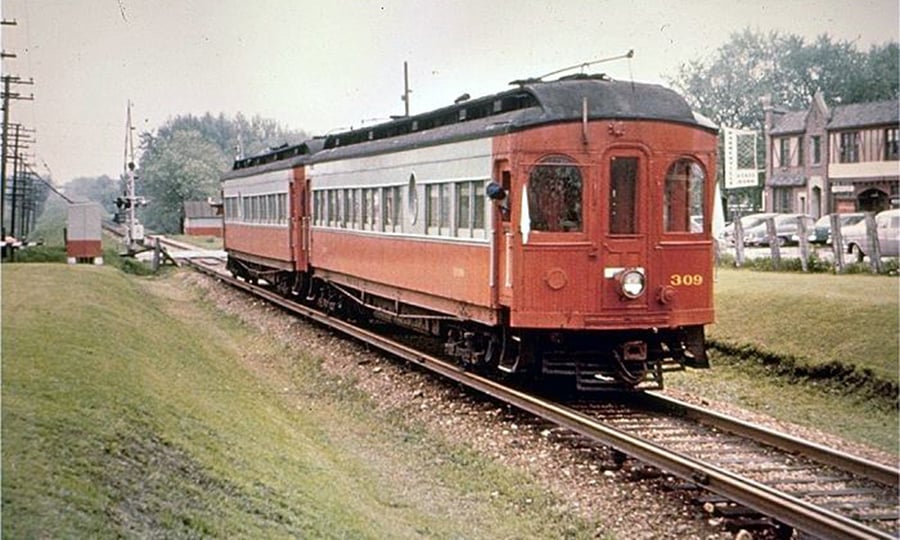 A red, two-car train travels past a small downtown area.