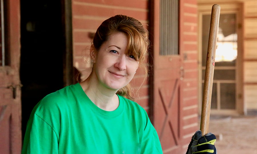 A woman in a green t-shirt stands outside of a horse stable wearing work gloves.