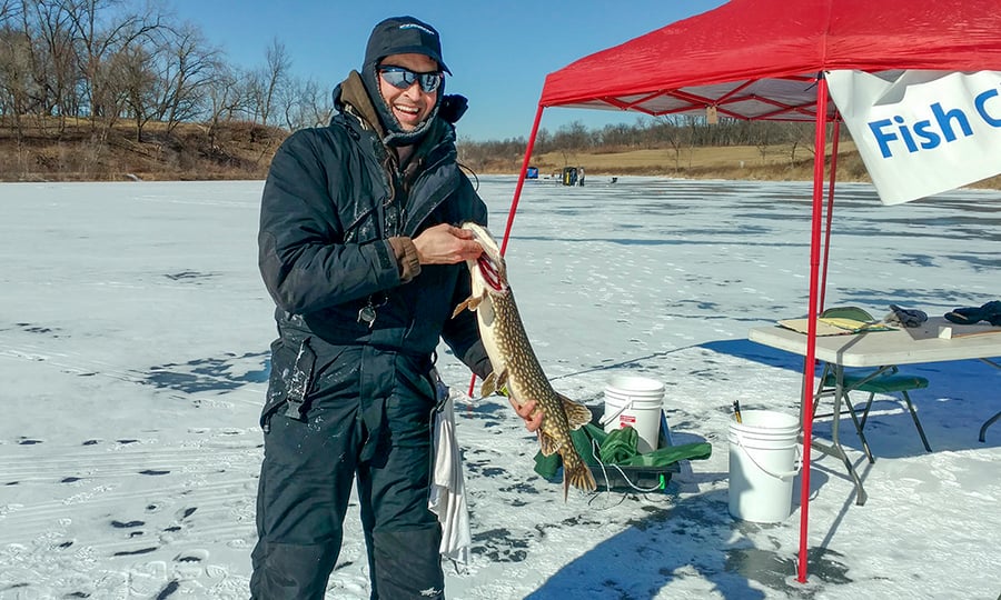 A man in a black snowsuit holds a northern pike while standing on a frozen lake.