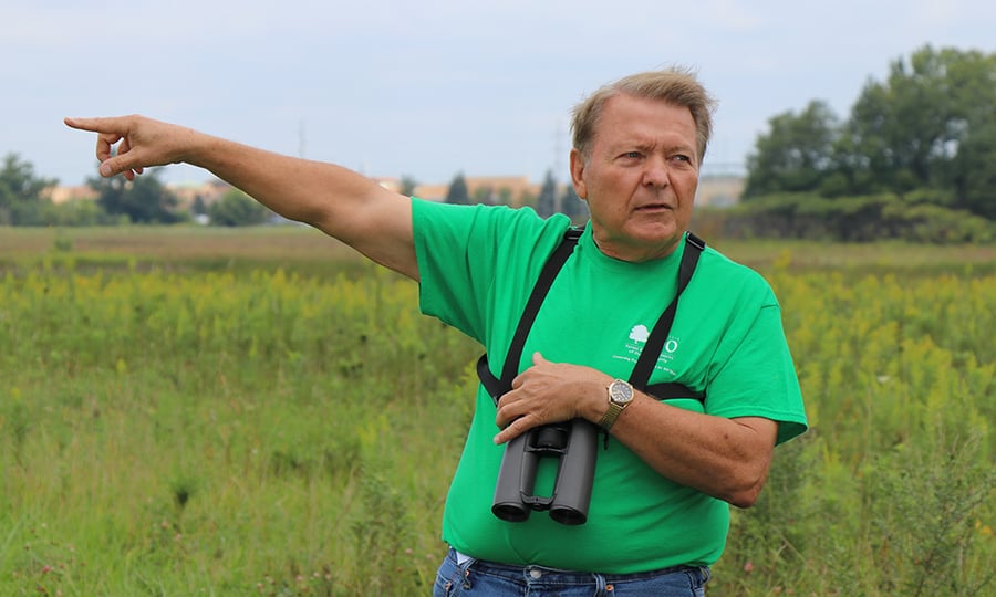 A man in a green shirt with binoculars around his neck points across a field.