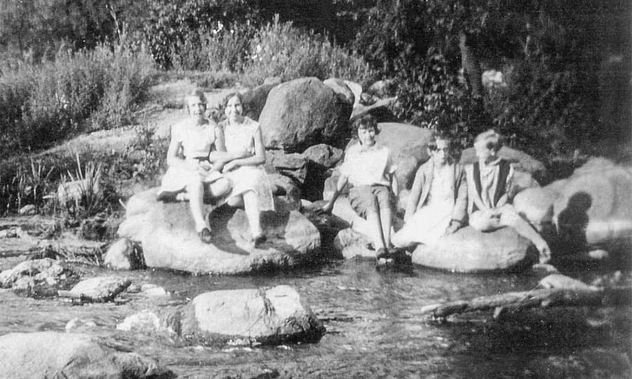 Five women sit on rocks on the side of a river in this old photograph.