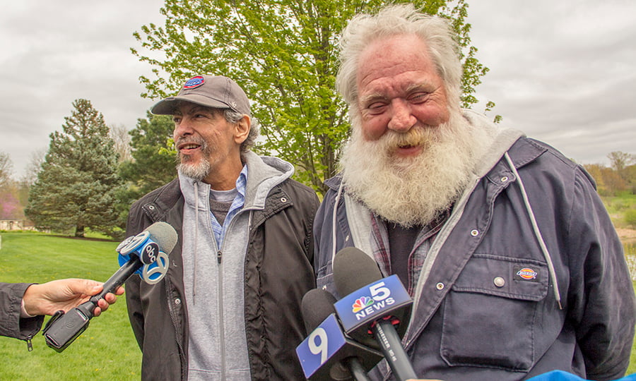 Two men talk to TV reporters in a grassy field.