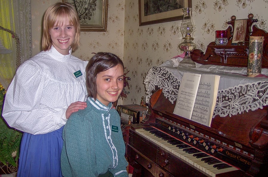 Two girls in 1880s attire pose for a photo in front of the organ.