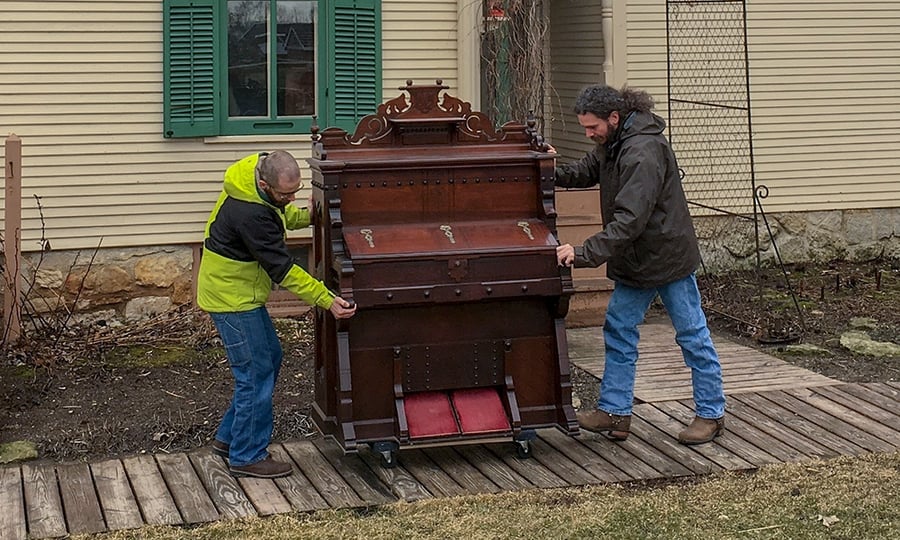 Two men roll a pipe organ down a wooden walkway.