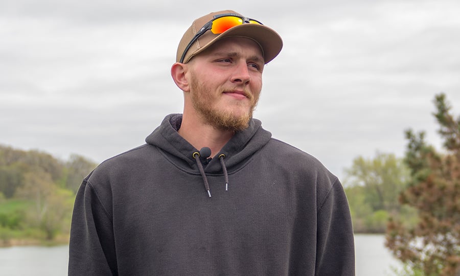 A man in a hat and sunglasses stands in front of a lake.