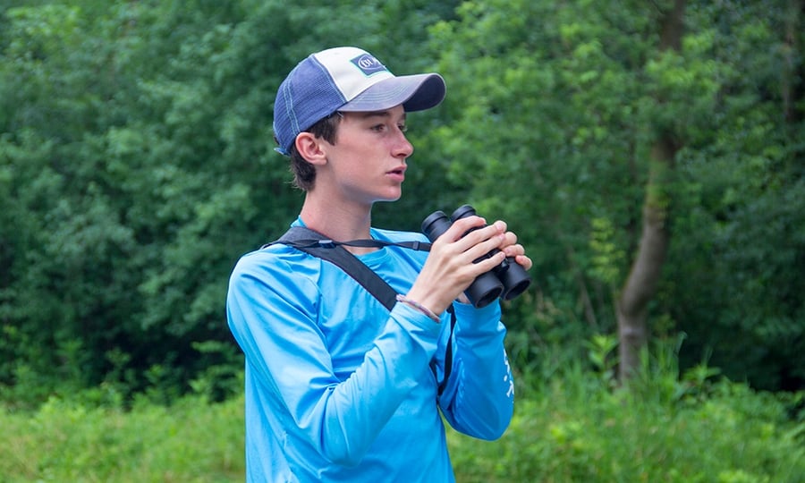 A guy in a hat and blue shirt holds binoculars while looking for birds on the edge of a forest.