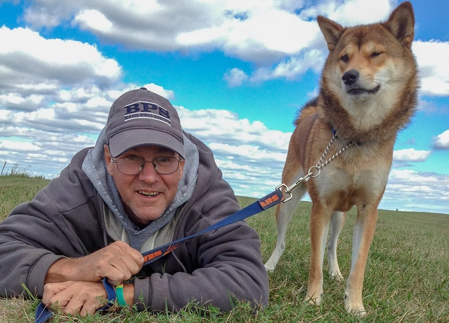 A man lays in a grassy field with his dog standing next to him under a partially cloudy blue sky.