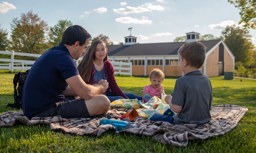 A family enjoys a picnic on a blanket in front of a horse barn.