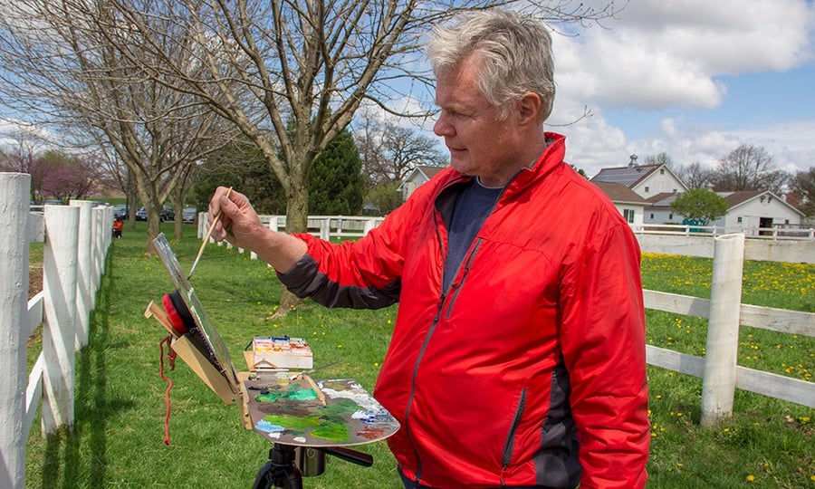 A man stands outside, painting on an easel.