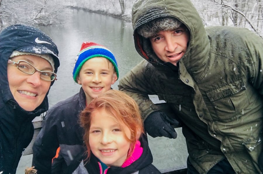 A family of four appropriately dressed in warm clothes pause for photo while it snows on the forest behind them.
