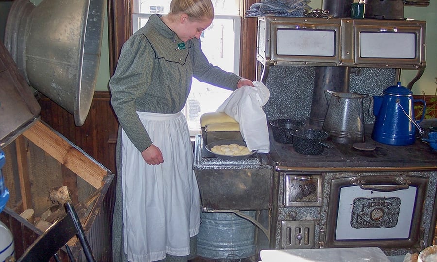 A woman uncloaks a pan of biscuits that sit atop a wood burning stove.