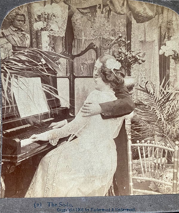 A man hugs a woman while their hands rest on a piano in this old photograph.