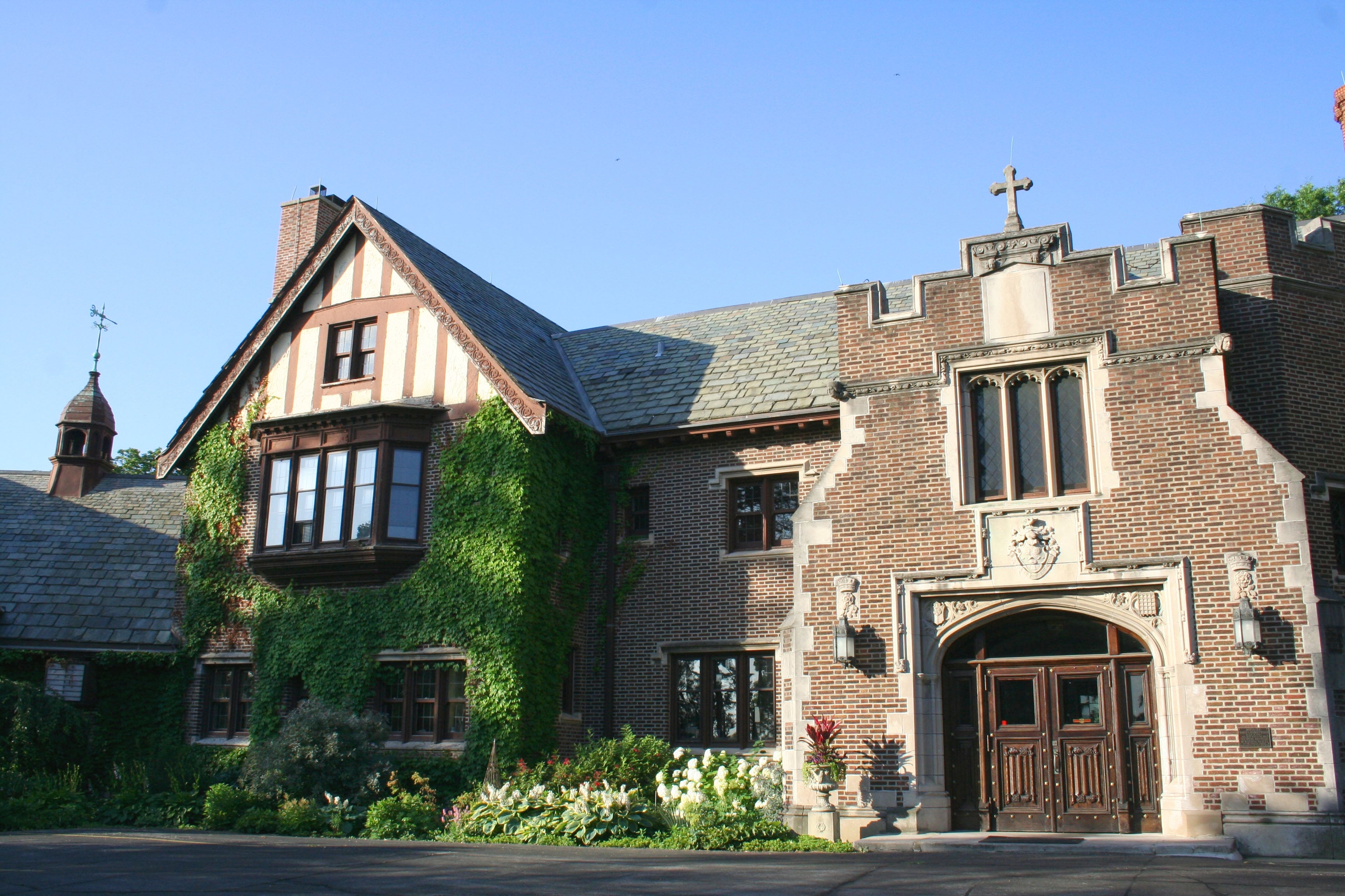 A Tudor-style mansion stands in sunlight with a blooming garden out front.