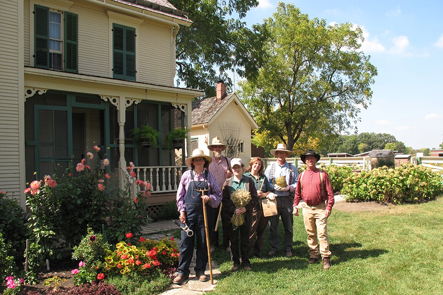 A group in 1880s attire stand in front of an old farmhouse.