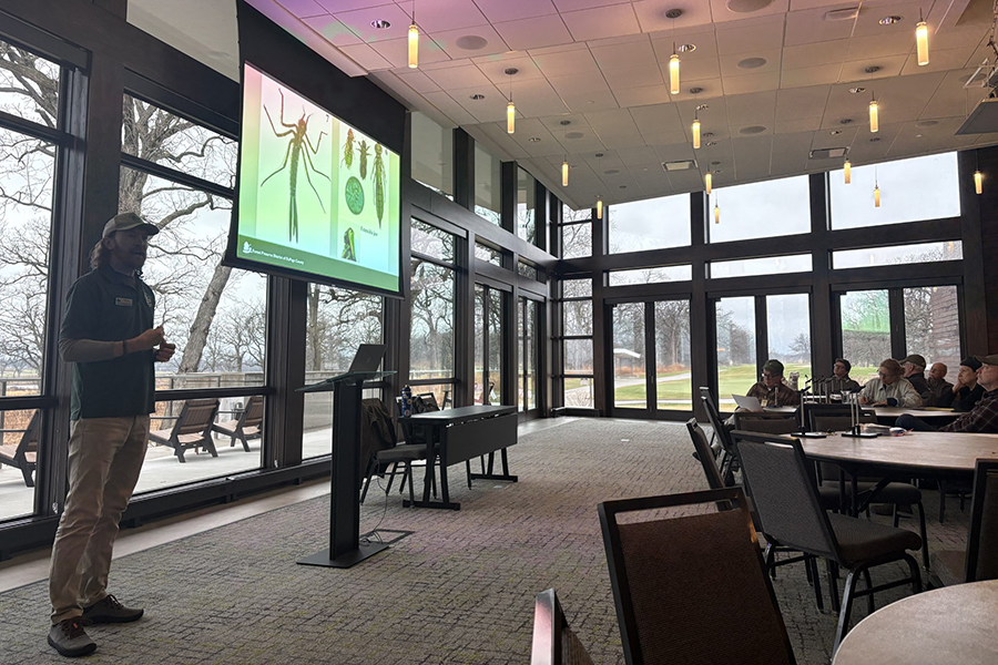 A man delivers a Powerpoint presentation in an event space surrounded by windows.