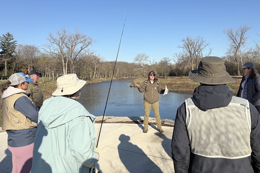 A woman speaks to a group while standing on a concrete platform above a creek on a chilly, sunny morning.