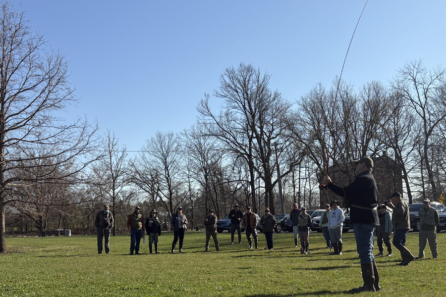 A man demonstrates how to cast a fly rod in a grassy field with about a dozen people looking on.