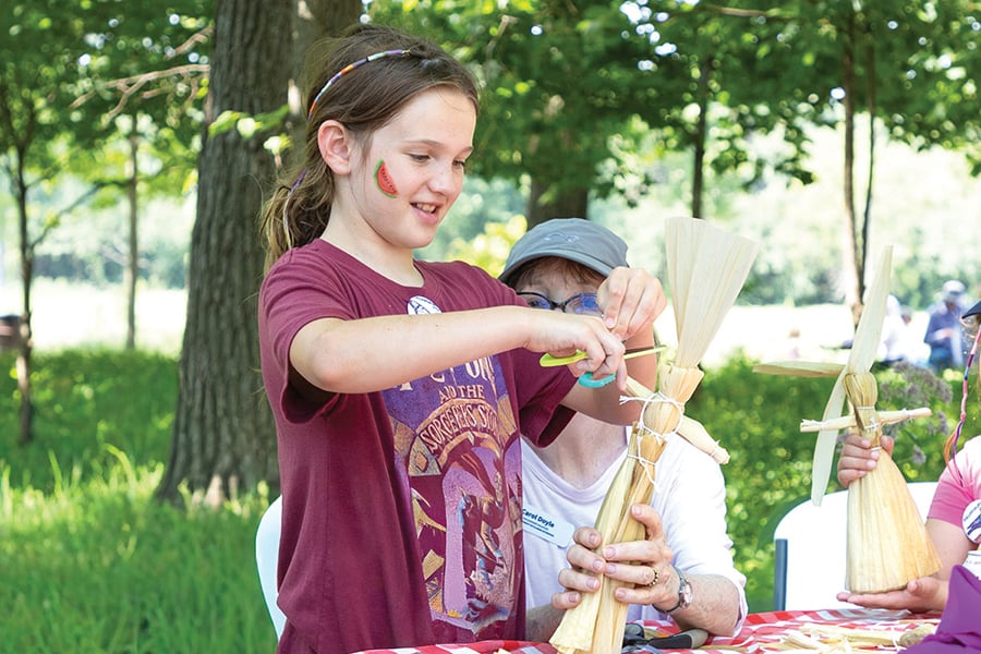 a girl making a corn husk doll at a table outside