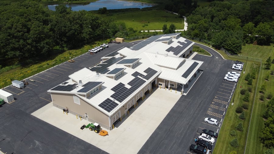 Solar panels rest atop a light-colored building surrounded by trees and a pond.