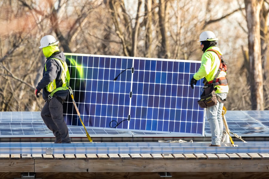 Two workers in neon tops carry a solar panel on top of a roof during installation.