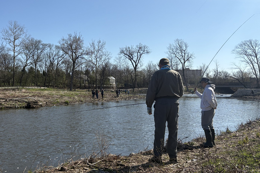A man gives pointers to a teen fly-fishing from the bank of a stream.